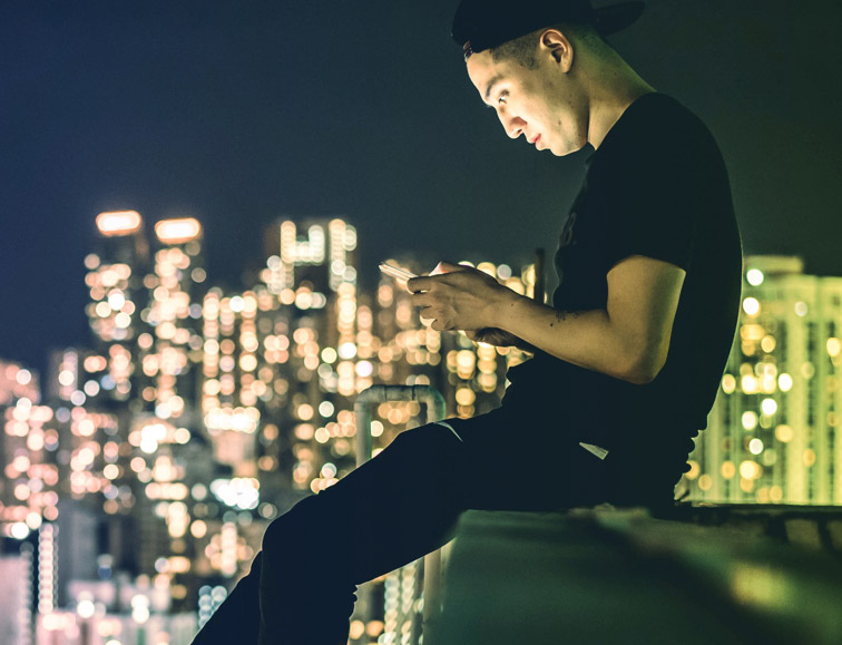 Image of a man sitting on the edge of a roof top on his phone with city skyline in the background.